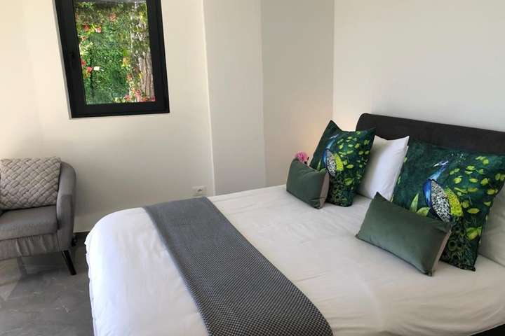 Stylish bedroom at Villa Cap offering a cozy grey armchair, vibrant peacock pillows, and a window framing lush greenery.