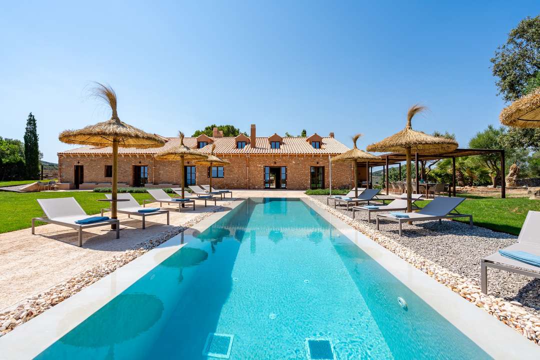 Luxury saltwater pool at Villa Medio, featuring sun loungers, straw parasols, and a stunning rustic stone facade in Mallorca.