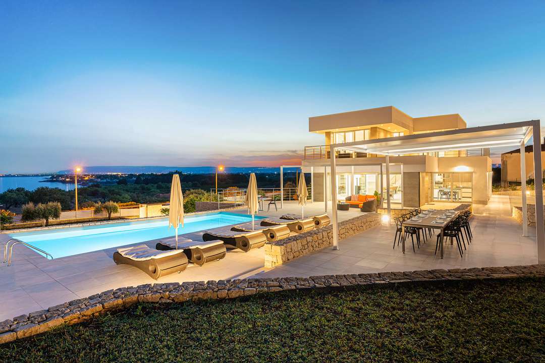 Contemporary Villa Nina at dusk, featuring a heated saltwater pool, elegant alfresco dining, and panoramic Mediterranean views.