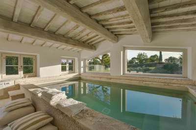 Luxurious indoor swimming pool with counter-current system, exposed beams, and large windows framing scenic Tuscan views.