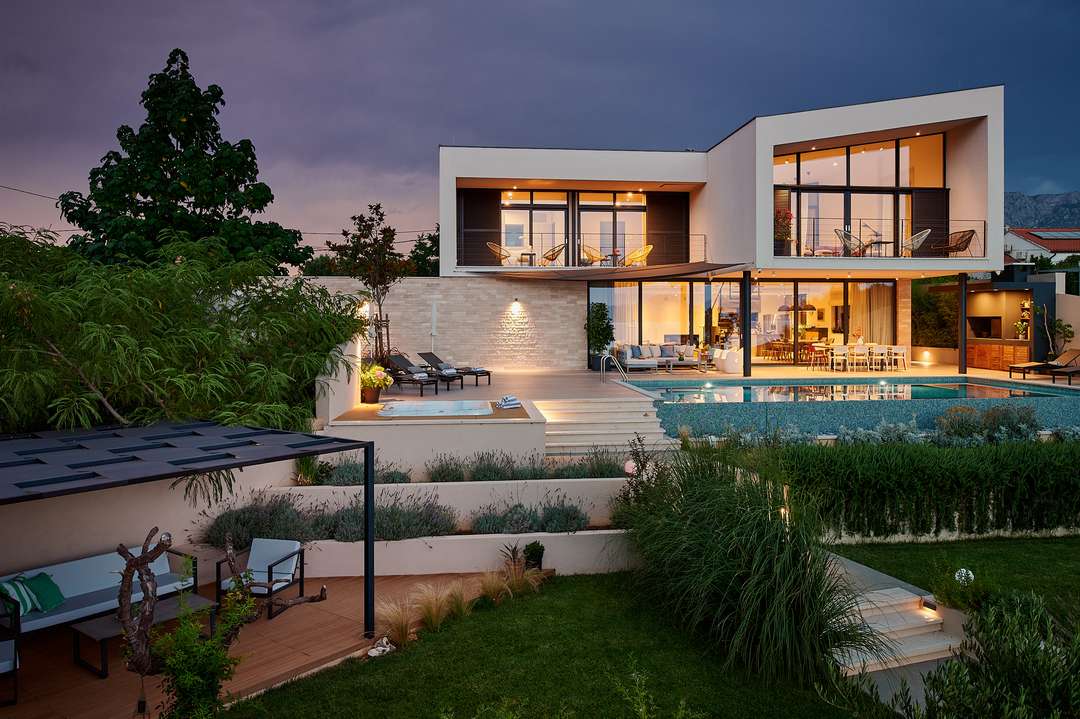 Evening view of Villa Pure Hvar's modern exterior, featuring a glowing heated pool, Jacuzzi, and elegant outdoor dining.