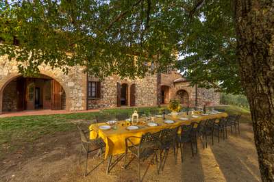 Elegant al fresco outdoor dining area under shaded trees beside the rustic stone facade of Villa Serenzia in Tuscany.